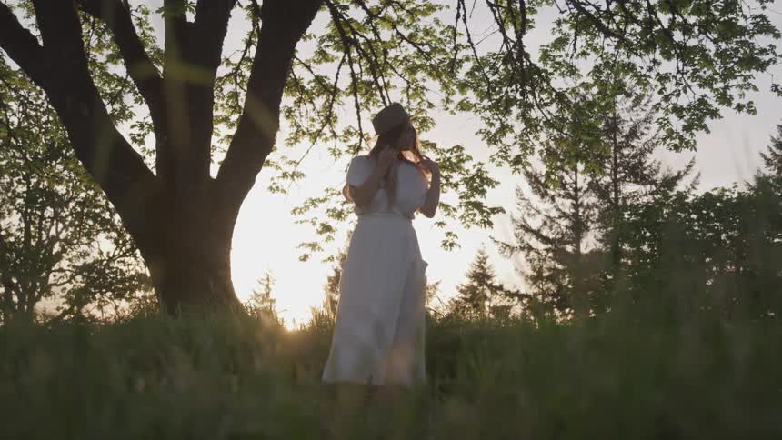 Blonde woman in white dress stands under a tree while the sun goes down