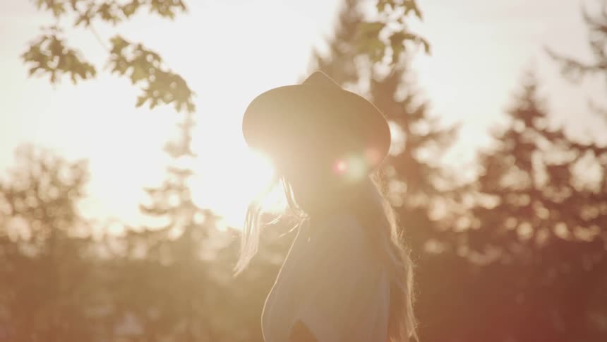 Woman wearing wide hat turns her head over her shoulders and smiles at sunset
