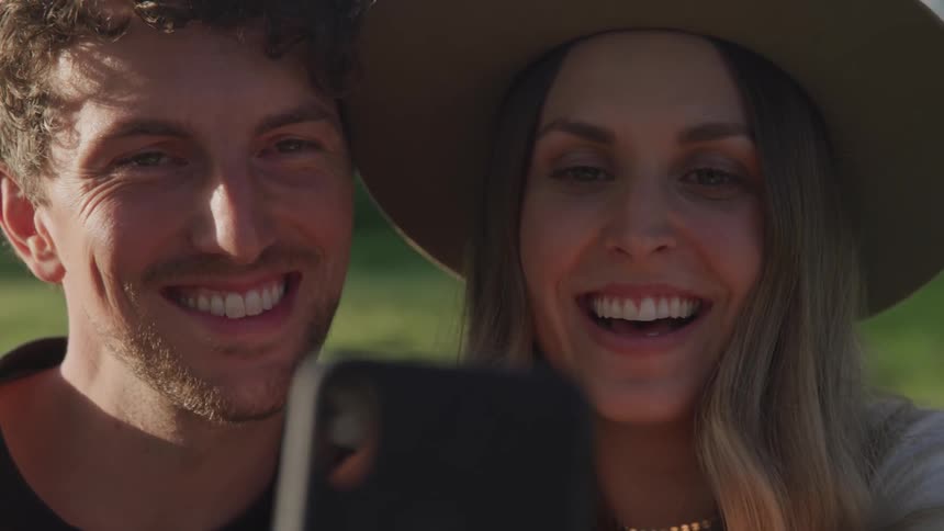 A man and woman side by side smiling as they take a selfie outside with grass behind them on a sunny day