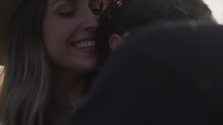A woman in a sun hat blowing the seeds of a dandelion into the face of a man