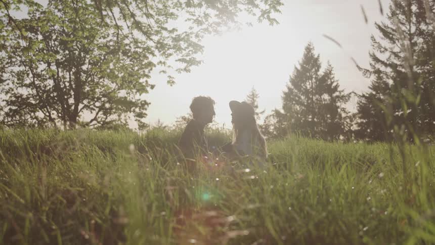 A young man and woman sit in a field holding hands and looking at one another