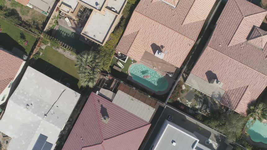 A woman is swimming in a clear blue pool in the small back yard of a house