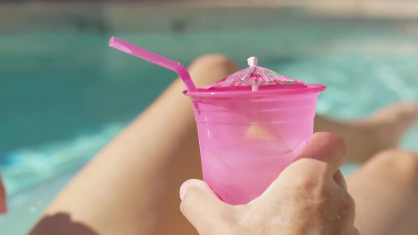 A woman drinks from a pink cup with a drink umbrella while floating in a swimming pool