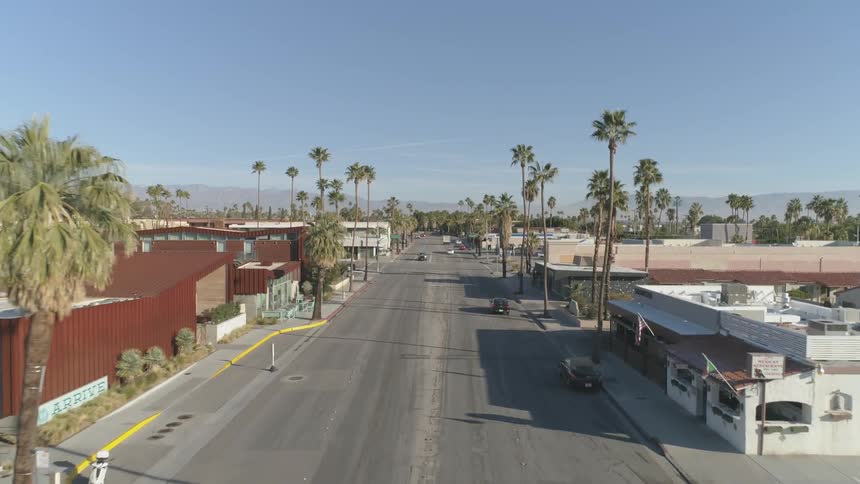 Cars drive along road between low buildings and palm trees