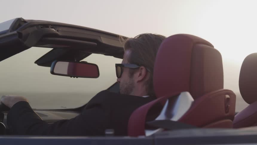 Blonde man wearing sunglasses and in suit drives convertible car at dusk