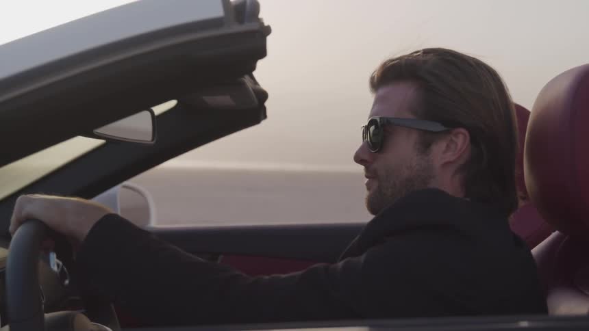 A man in a jacket is driving a convertible with the top down and his sunglasses on