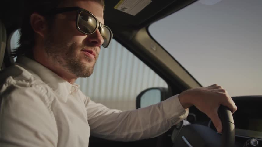 A man wearing sunglasses looks around as he drives a car
