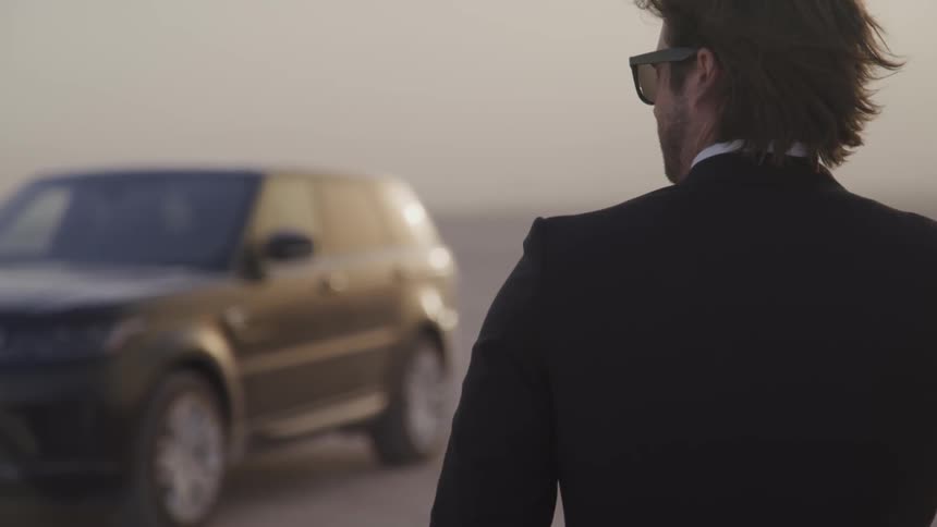 A man in a suit is looking at a parked range rover while his hair blows in the wind