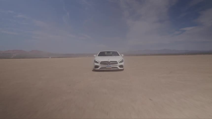 A white mercedes benz is driving at a fast speed through the desert on a partly cloudy day