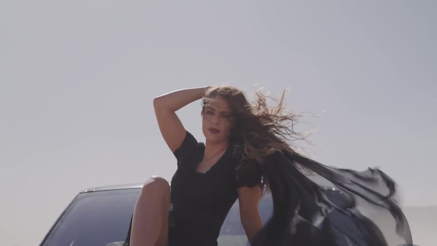 A woman sits on the hood of a car with her clothes and hair blowing in the wind