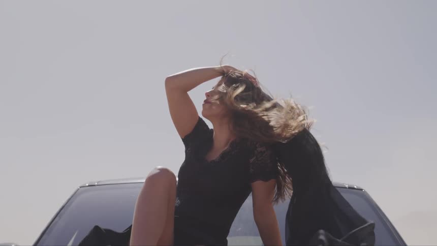 A woman sits on the hood of a car while her hair blows in the wind