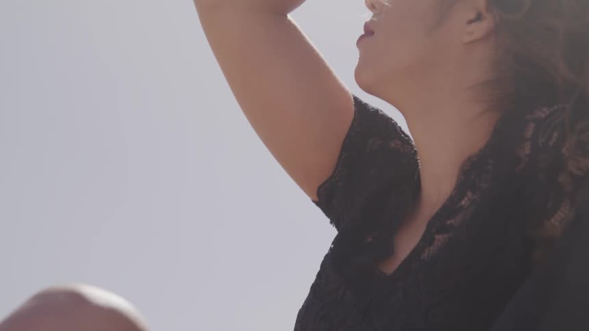 A woman sits with her hand on her head as her black shirt and brown hair billow in the breeze