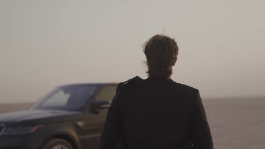 A man in a windy desert walks to an suv