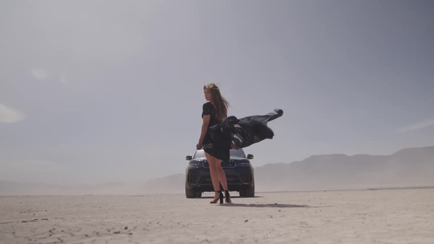 A woman stand in the sand in front of a car while her dress blows in the wind