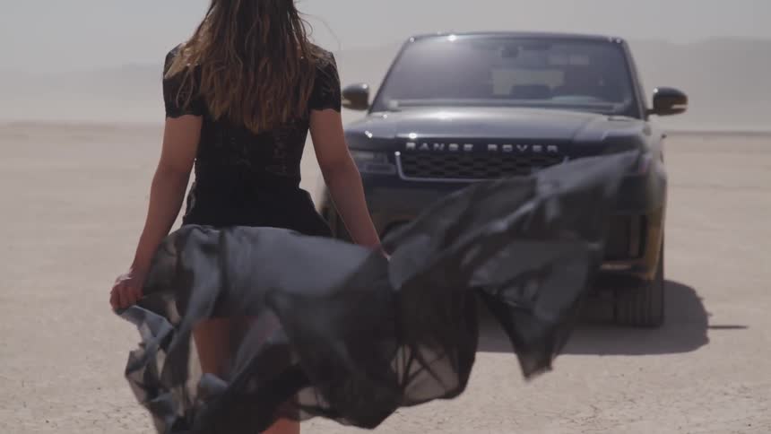 A woman stands in front of a vehicle with her dress blowing in the wind