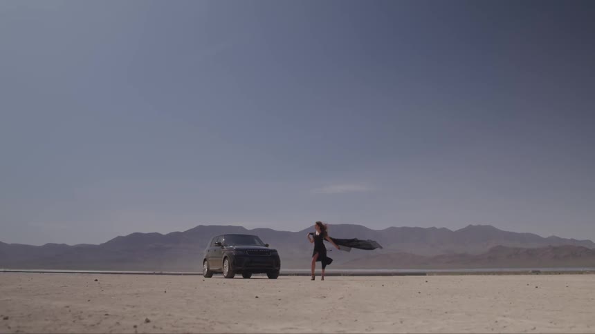 A woman with a black dress and scarf blowing in the wind in the desert next to a black truck