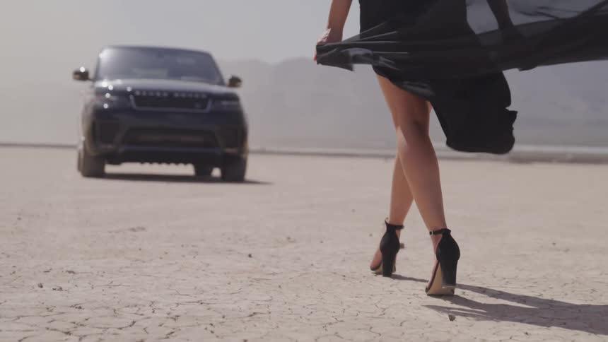 A woman wearing heels and a dress walks towards a vehicle in the desert