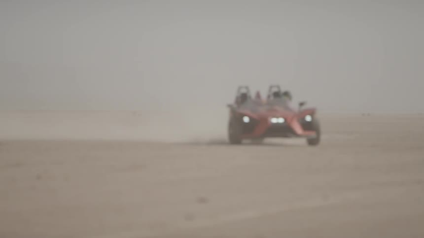 A red and black polaris slingshot races over desert sand leaving a dust trail