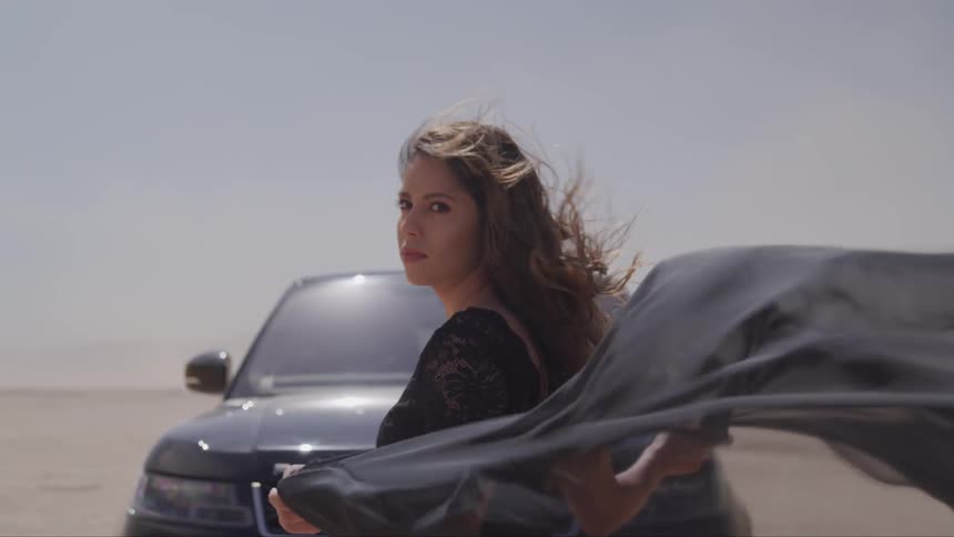 A woman stands in front of a car holding a billowing scarf