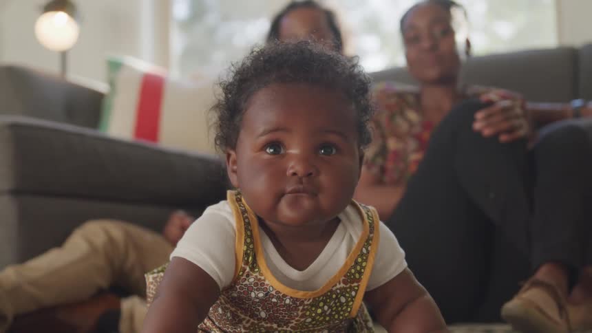 Mother and father are sitting on the floor watching their baby with pride as she holds her head up while laying on her stomach and then the baby sneezes