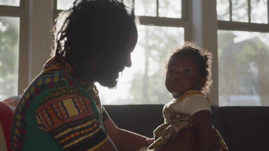A man in a living room sitting on a sofa talking to a baby in his lap