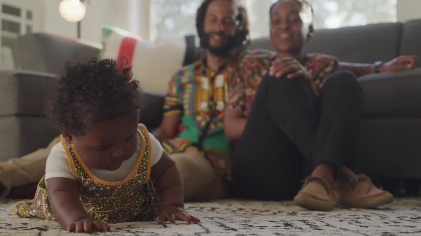A baby plays on a black and white rug while her mom and dad sit on the floor behind her