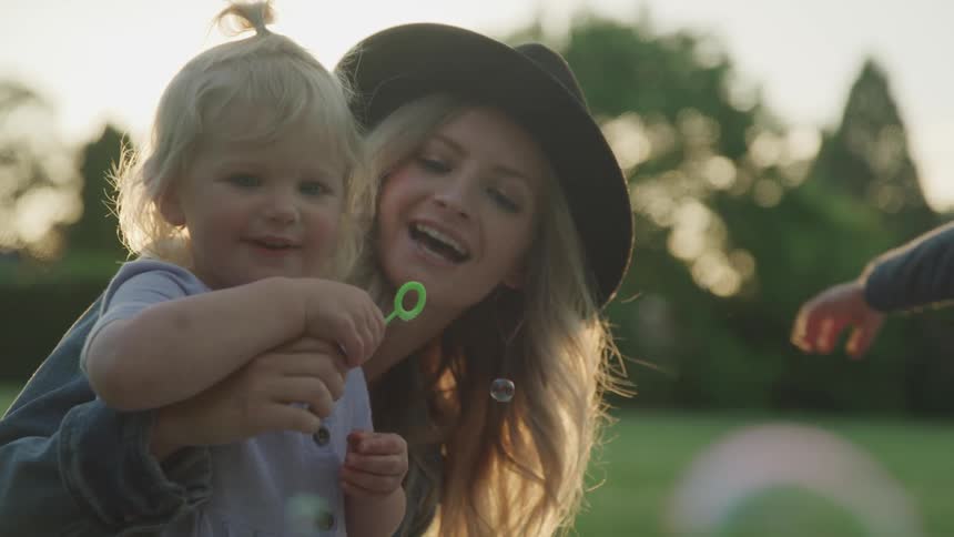 Blonde mother play bubbles with her daughter at public park on the sunset
