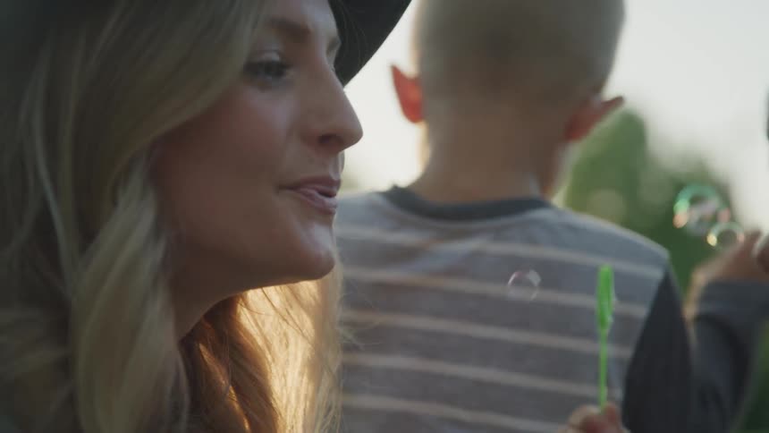 A blonde white woman blows bubbles outdoors with her children