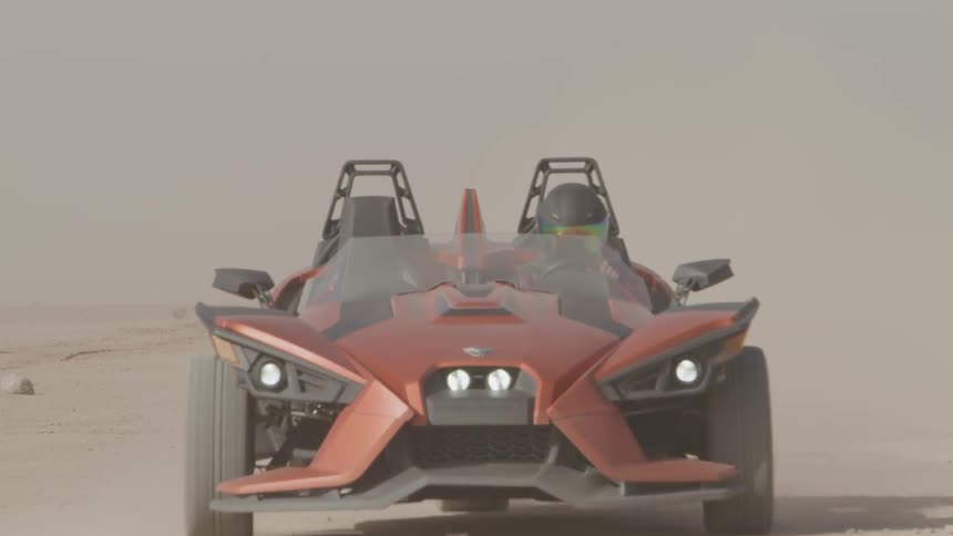 A person wearing a helmet drives a red and black three wheeled vehicle kicking up dust