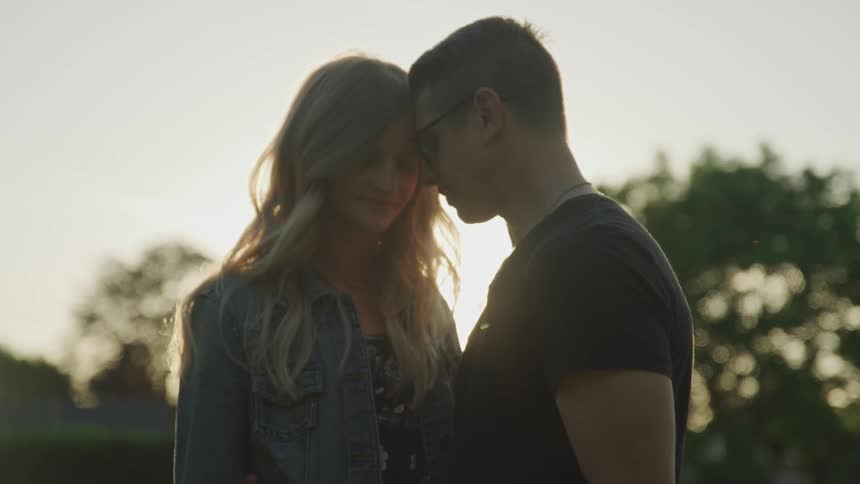 A woman and man standing outdoors pressing their foreheads together at sunset