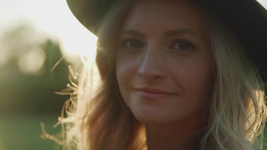 Blonde woman wearing wide hat looks at the camera and smiles at sunset