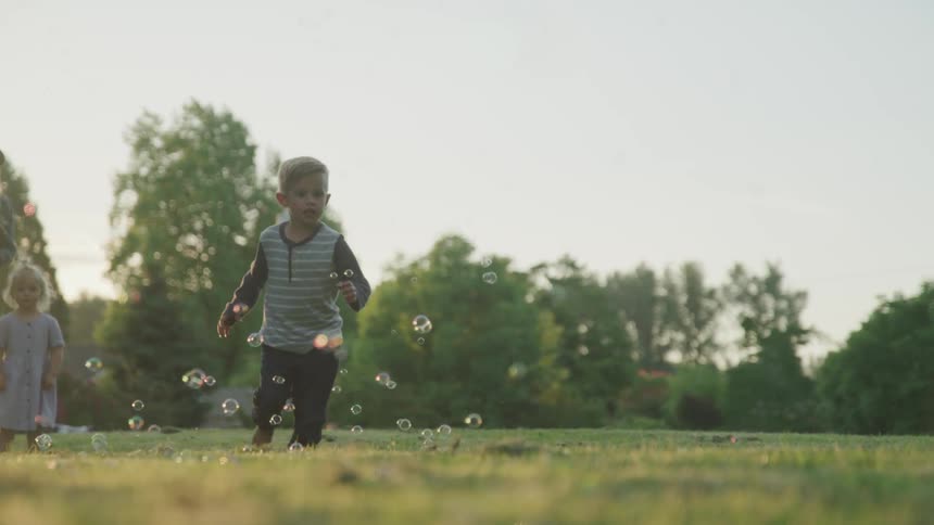 A young boy runs around trying to catch bubbles floating in the air while a girl watches in the background