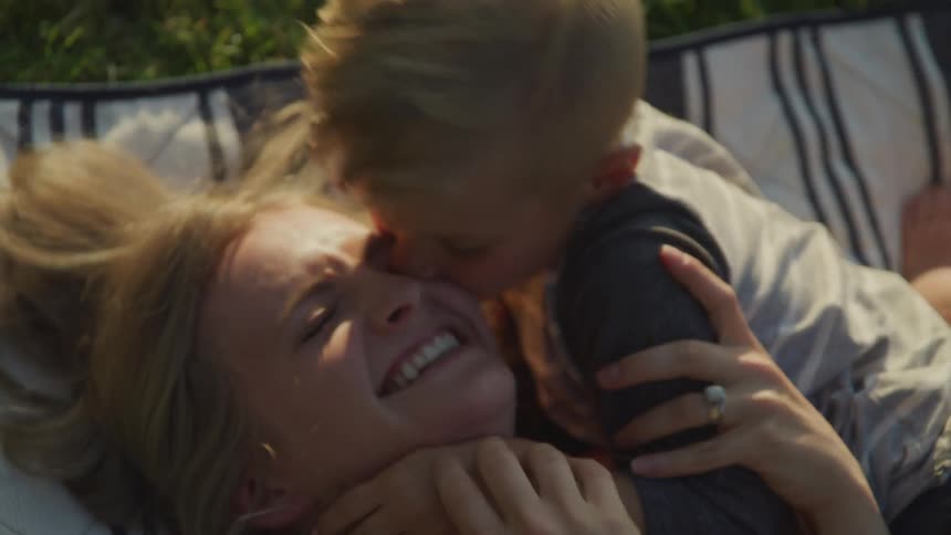 A young boy is hugging and kissing his moms face as they lay on a blanket in the grass