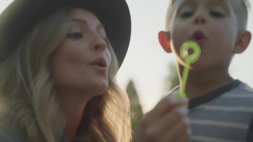 A woman and a young boy are blowing bubbles through a bubble wand that the boy then tries to catch