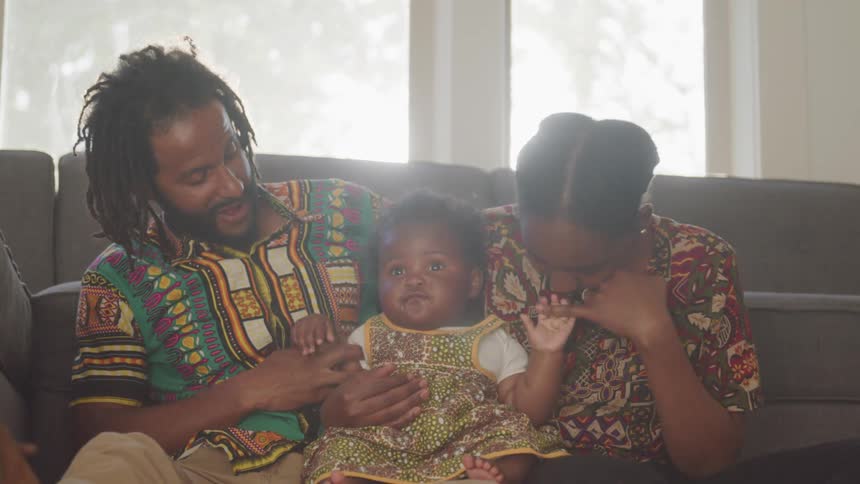 A black man and woman sit on the floor in front of a couch and play with the hands of their chubby baby who is wearing a dress