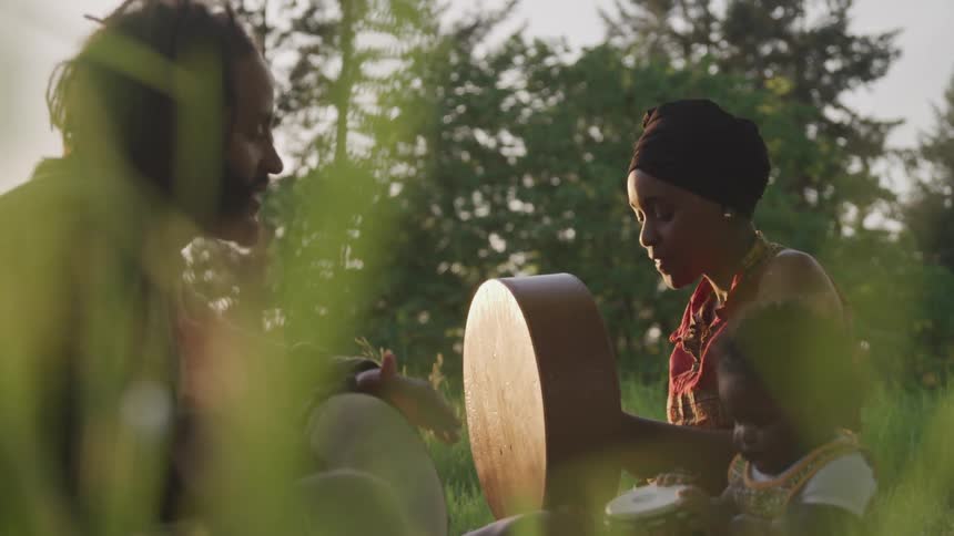 Man in white shirt is playing a drum while a woman in a red dress wearing a brown scarf on her head watches her small child playing on its own drum