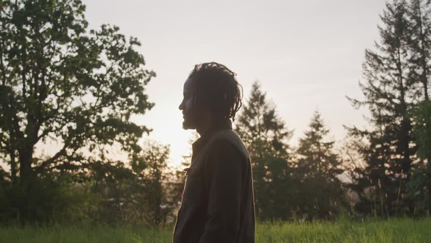 A man with disheveled hair faces sideways as he stands in a grassy field near some deciduous and evergreen trees