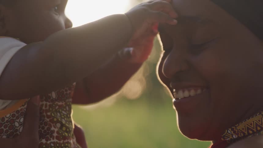 A woman holds her baby up in the sunshine outdoors and has a huge smile on her face