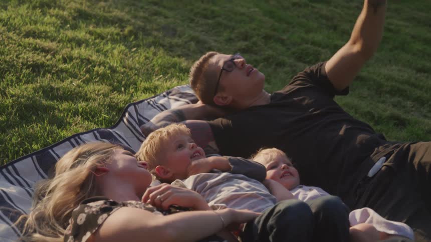 A young family smiles and laughs while lying on a blanket on a grassy lawn