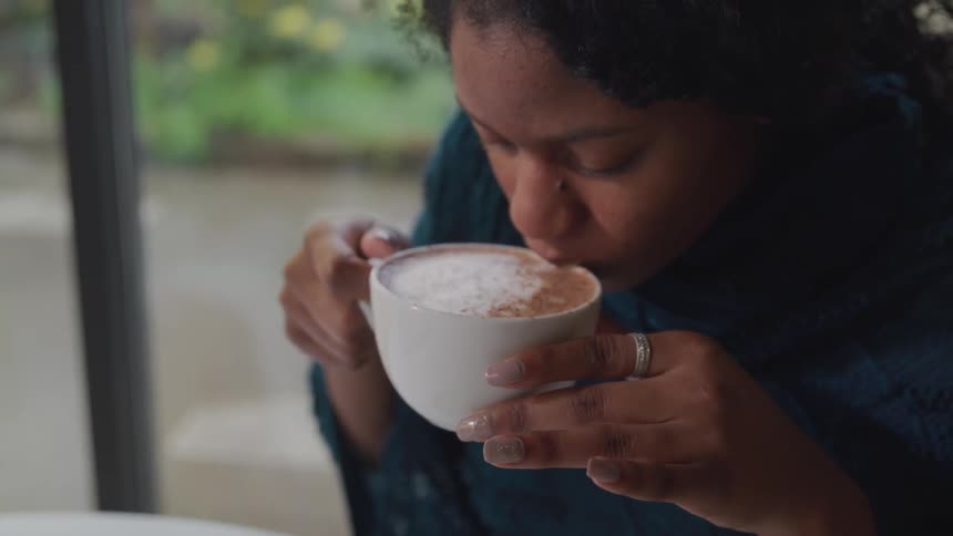 A woman drinks a cappuccino from a white mug while sitting at a white table