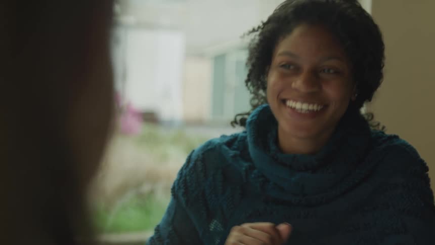 A young black woman smiles and laughs as she sits indoors and converses with another person