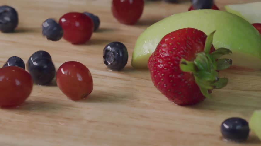Blueberries strawberries and apple slices are dropped onto a wood grain counter top