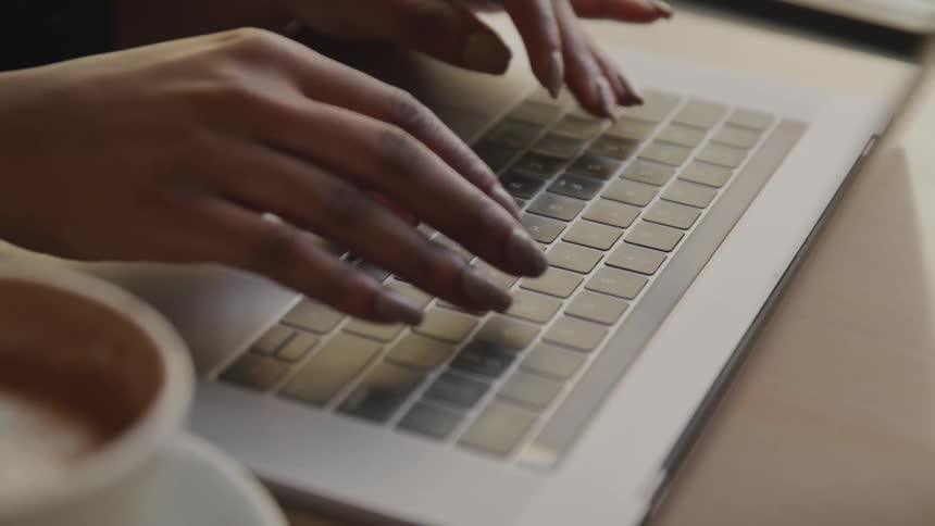A woman with long fingernails is typing carefully on a laptop keyboard with a cup of coffee next to her