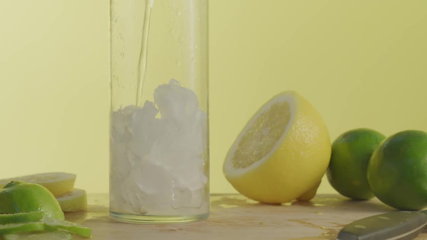 Lemons and limes surround a glass of ice being filled with liquid