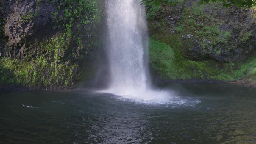 Water from a waterfall is splashing down from a great height into a body of water surrounded by rocky walls covered in green foliage
