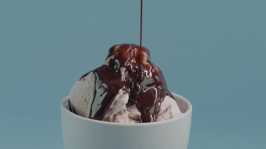 Chocolate source poured on ice cream in a bowl set against a blue studio background