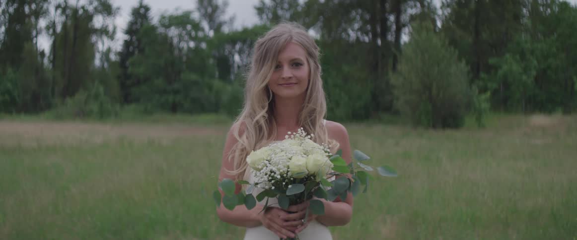 Female with long blonde hair is standing in a field of tall grass wearing a white dress and holding a bouquet of white roses