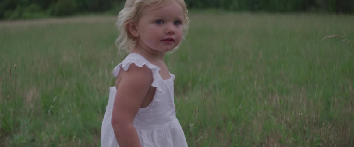 Blonde blue eyed toddler girl walks on the grass on a cloudy day
