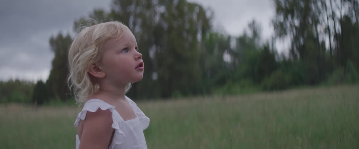 Blonde blue eyed toddler girl standson the grass and looks up in the field on a cloudy day