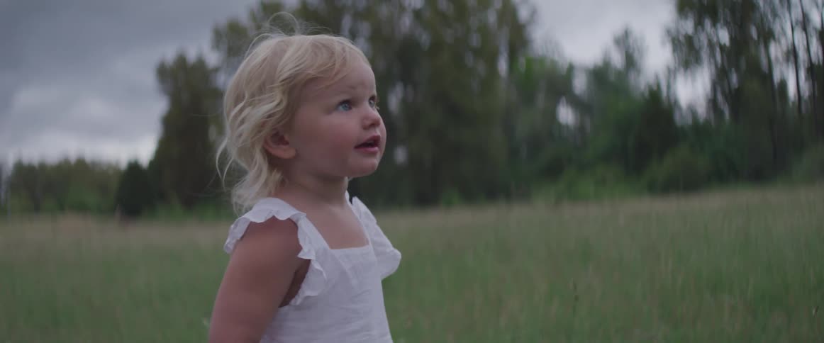 Blonde blue eyed toddler girl looks up on the grass on a cloudy day